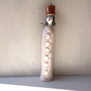 May include: A ceramic figurine of a person wearing a brown crown and a long, white dress with white buttons. The figurine has a painted face with black hair and red lips.