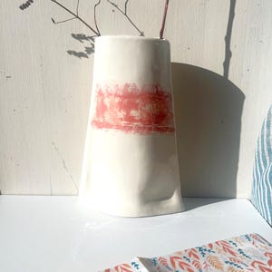 May include: White ceramic vase with a pink and orange abstract design. The vase is tall and narrow with a slightly curved shape.
