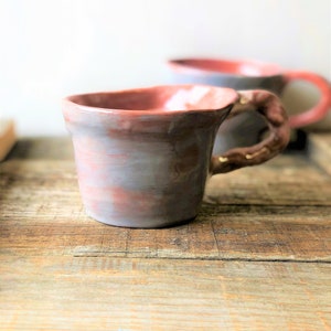 May include: Two handmade ceramic mugs with a light gray and pink glaze. The mugs have a unique, slightly uneven shape and a textured finish.