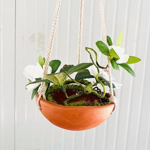 May include: A hanging terracotta planter with white flowers and green leaves. The planter is suspended by beige rope and filled with greenery and white blossoms. The pot has a rounded shape and is filled with green moss.
