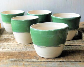 May include: Five ceramic mugs with a green and white glaze. The mugs are arranged in a row on a wooden surface.