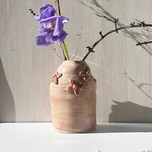 May include: A light pink ceramic vase with a textured surface and brown flower accents. A single purple flower is in the vase.
