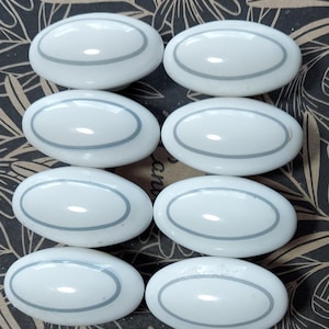 May include: Eight white oval cabinet knobs with a gray border. The knobs are arranged on a patterned background. These knobs are perfect for adding a touch of elegance to any furniture piece.