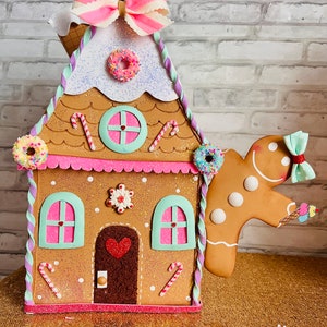 May include: A gingerbread house decoration with a pink and white bow on top, a gingerbread man with an ice cream cone, and candy decorations. The house is made of brown, pink, and white felt and has a white glitter roof. The gingerbread man is made of brown felt and has a white icing smile.