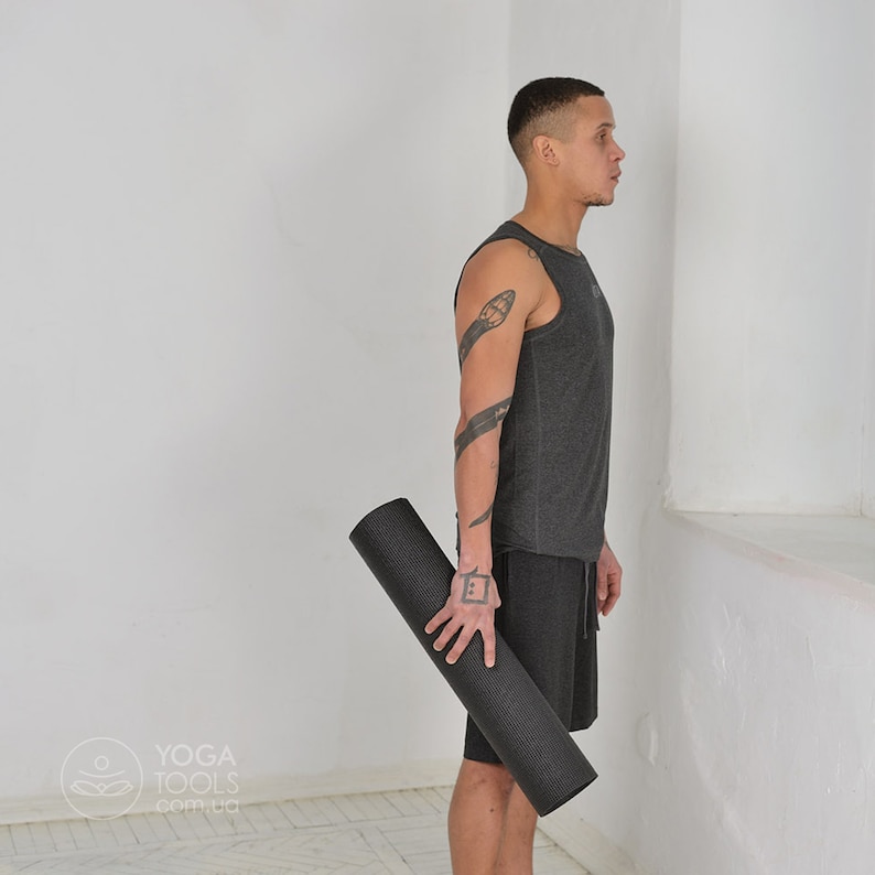 K&ouml;nnte beinhalten: Eine Person h&auml;lt eine schwarze Yogamatte. Die Matte ist zusammengerollt und hat eine strukturierte Oberfl&auml;che. Die Person tr&auml;gt ein graues Tanktop und schwarze Shorts.