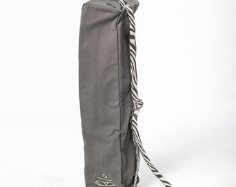 Puede incluir: Una bolsa de yoga gris con una correa de estampado de cebra. La bolsa tiene un cierre de cremallera y un logotipo en la parte delantera.