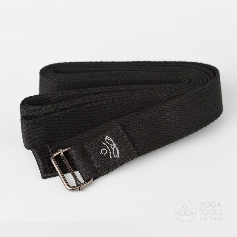 May include: Black yoga strap with a silver buckle and a small logo embroidered on the strap. The strap is rolled up and the buckle is visible.