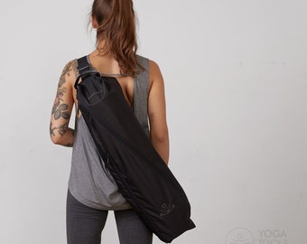 Puede incluir: Una bolsa negra para esterilla de yoga con correa para el hombro. La bolsa es larga y cilíndrica, diseñada para transportar una esterilla de yoga enrollada. La bolsa tiene cierre de cremallera y un pequeño logotipo en el lateral. La persona lleva una camiseta sin mangas gris y leggings.