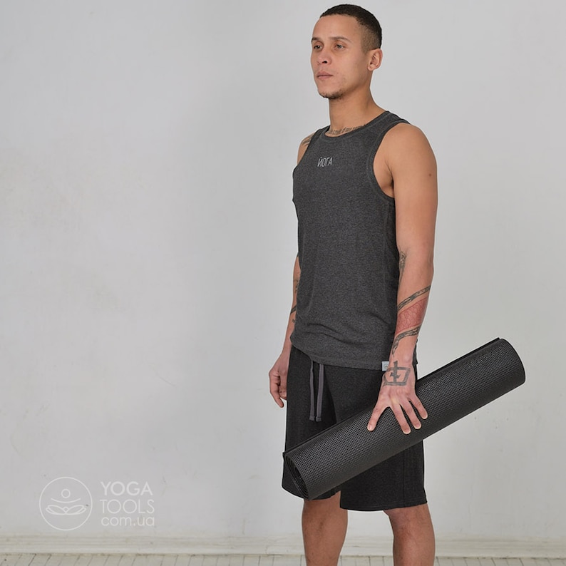 K&ouml;nnte beinhalten: Eine Person tr&auml;gt ein graues Tanktop mit dem Wort "Yoga" auf der Vorderseite, schwarze Shorts und h&auml;lt eine schwarze Yogamatte.