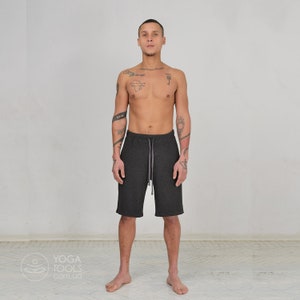 K&ouml;nnte beinhalten: Ein Mann tr&auml;gt dunkelgraue Sport-Shorts mit einem Kordelzug in der Taille. Er steht mit leicht ge&ouml;ffneten Beinen und die Arme h&auml;ngen an seinen Seiten. Er hat T&auml;towierungen auf seiner Brust, seinen Armen und seinem Hals.