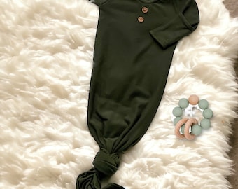 May include: A dark green knotted baby gown with long sleeves and three wooden buttons. The gown is lying on a white fluffy surface.