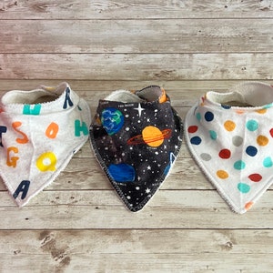 Könnte beinhalten: Drei weiße Baumwoll-Babyspucktücher mit verschiedenen Mustern. Ein Spucktuch hat ein buntes Alphabetmuster, ein anderes ein Weltraum-Muster mit Planeten und Sternen und das dritte ein buntes Punkte-Muster.