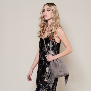 May include: A gray suede bucket bag with a fringe trim and a drawstring closure. The bag is worn by a person with long blonde hair and a floral dress.