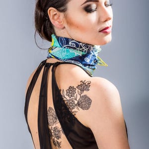 May include: A woman wearing a black sleeveless dress with a floral tattoo on her shoulder. She is wearing a colourful patterned scarf around her neck.