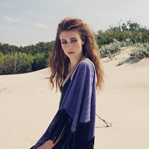 May include: A woman wearing a blue suede fringed shawl over a black top. The shawl features a perforated pattern and black fringe. The background shows a sandy beach and green vegetation.