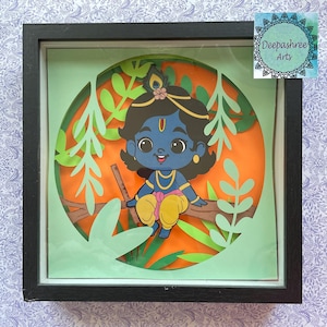 May include: A framed paper art piece depicting a cartoon image of Krishna, a Hindu deity, sitting on a branch with green leaves. The artwork is in a black frame and has a green and orange background.