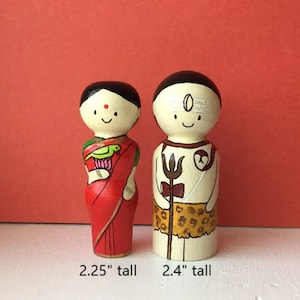May include: Two hand-painted wooden dolls, one wearing a red sari with a green parrot and the other wearing a white tunic with a brown animal print skirt and holding a trident. The dolls are 2.25 inches and 2.4 inches tall.