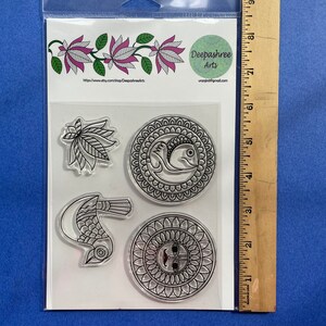 May include: A clear plastic sheet with four clear stamps featuring black and white designs. The designs include a bird, a fish, a floral pattern, and a sun with a face. The sheet is packaged in a clear plastic bag with a white label that reads "Deepashree Arts" and a website address.
