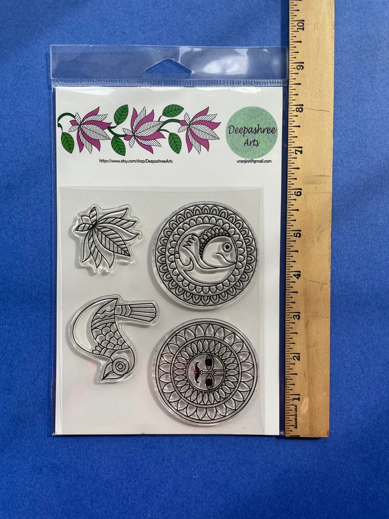 May include: A clear plastic sheet with four clear stamps featuring black and white designs. The designs include a bird, a fish, a floral pattern, and a sun with a face. The sheet is packaged in a clear plastic bag with a white label that reads "Deepashree Arts" and a website address.