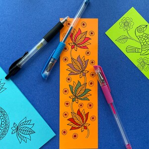 May include: An orange bookmark with a floral design featuring four stylized lotus flowers in shades of red, orange, green, and brown. The bookmark is surrounded by three pens: one black, one blue, and one pink.