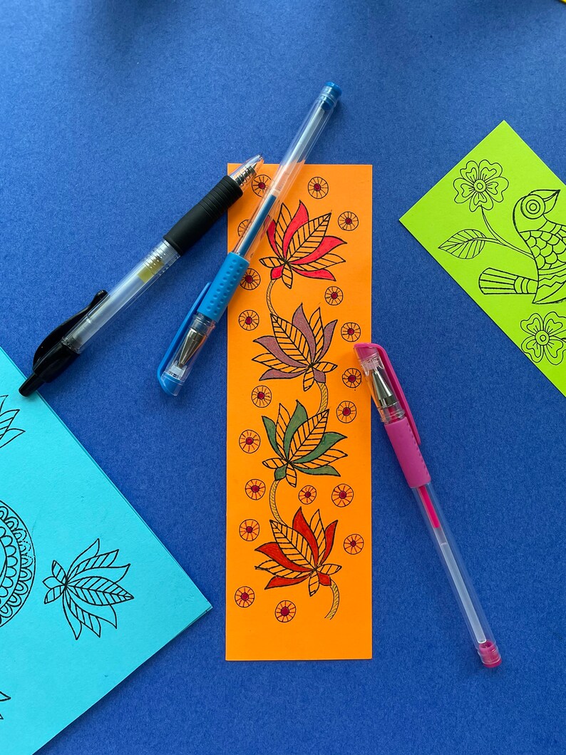 May include: An orange bookmark with a floral design featuring four stylized lotus flowers in shades of red, orange, green, and brown. The bookmark is surrounded by three pens: one black, one blue, and one pink.