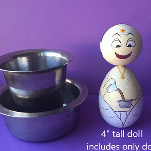 May include: A 4" tall wooden doll painted to look like a person pouring liquid from a white pitcher into a small metal cup. The doll is wearing a white robe with a gold necklace. The doll is standing on a purple surface.