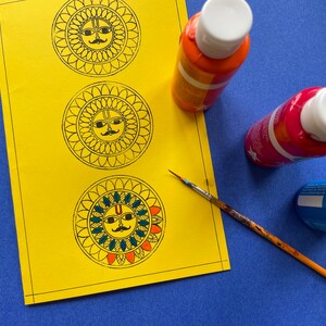 May include: A yellow sheet of paper with three black and white mandala designs featuring a sun face with a mustache.  There are three paint bottles, one orange, one red, and one blue, and a paintbrush on the paper.