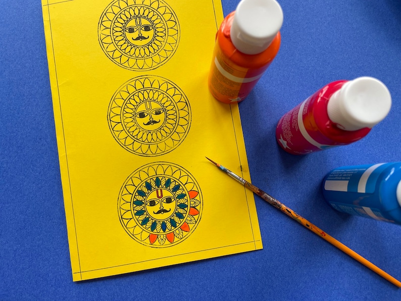 May include: A yellow sheet of paper with three black and white mandala designs featuring a sun face with a mustache.  There are three paint bottles, one orange, one red, and one blue, and a paintbrush on the paper.