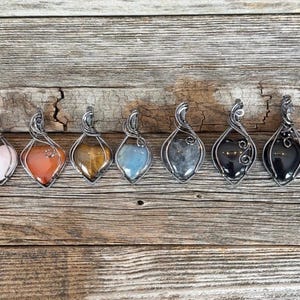 May include: A collection of seven gemstone pendants with silver wire wrapping. The gemstones include pink, orange, tiger's eye, blue, gray, black, and black. The pendants are displayed on a weathered wooden surface.