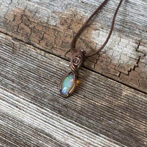 May include: A handcrafted pendant necklace featuring a wire-wrapped opal gemstone. The opal has a pearlescent, iridescent quality, set in a copper-colored metal. The necklace hangs from a brown cord, set against a rustic wooden background.