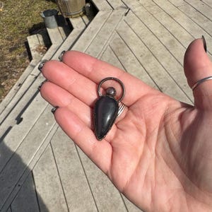 May include: A dark gray pendant with a teardrop-shaped stone, framed in silver metal. The pendant has a small silver ring at the top. The stone is smooth and polished, with a slight sheen. The pendant is held in a hand.
