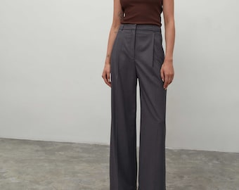 May include: A brown sleeveless ribbed tank top paired with high-waisted charcoal gray wide-leg trousers. The trousers feature a structured waistband with belt loops, a front zipper closure, and pressed creases running down the center of each leg.