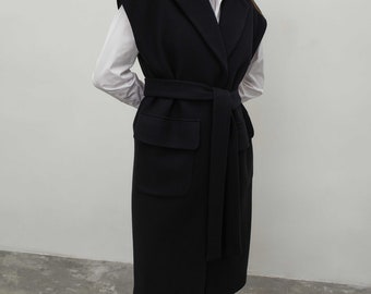 May include: A long black sleeveless vest featuring a wide lapel collar, a matching fabric tie belt at the waist, and two large flap pockets. The vest is worn over a crisp white collared shirt, paired with black trousers and black shoes.