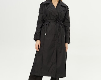 May include: A black trench coat featuring a wide collar, double-breasted button closure, and a matching fabric tie belt at the waist. The coat has long sleeves with buttoned cuffs and falls to a mid-calf length, paired with black trousers.