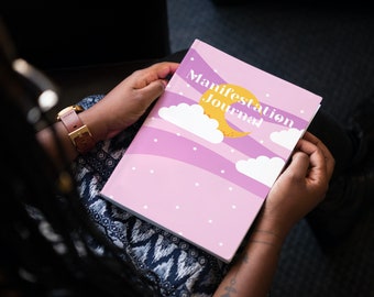 Può includere: Un diario di manifestazione rosa con una luna crescente gialla e nuvole bianche sulla copertina. Il testo "Manifestation Journal" è stampato in bianco.