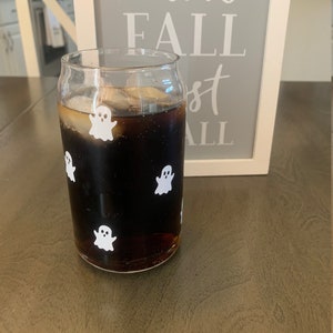 May include: A glass with a dark liquid and ice cubes, decorated with white ghost stickers. The glass is sitting on a wooden surface. The text "i love fall most of all" is visible on a sign behind the glass.