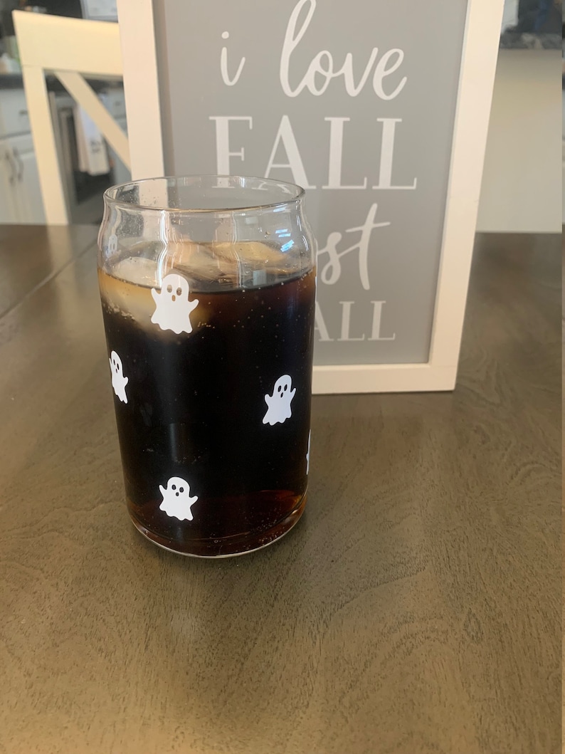 May include: A glass with a dark liquid and ice cubes, decorated with white ghost stickers. The glass is sitting on a wooden surface. The text "i love fall most of all" is visible on a sign behind the glass.