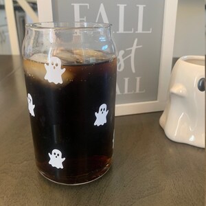May include: A glass of cola with ice and ghost stickers on the side. The glass is sitting on a wooden table next to a white ghost figurine and a framed sign that says "I love fall most of all".