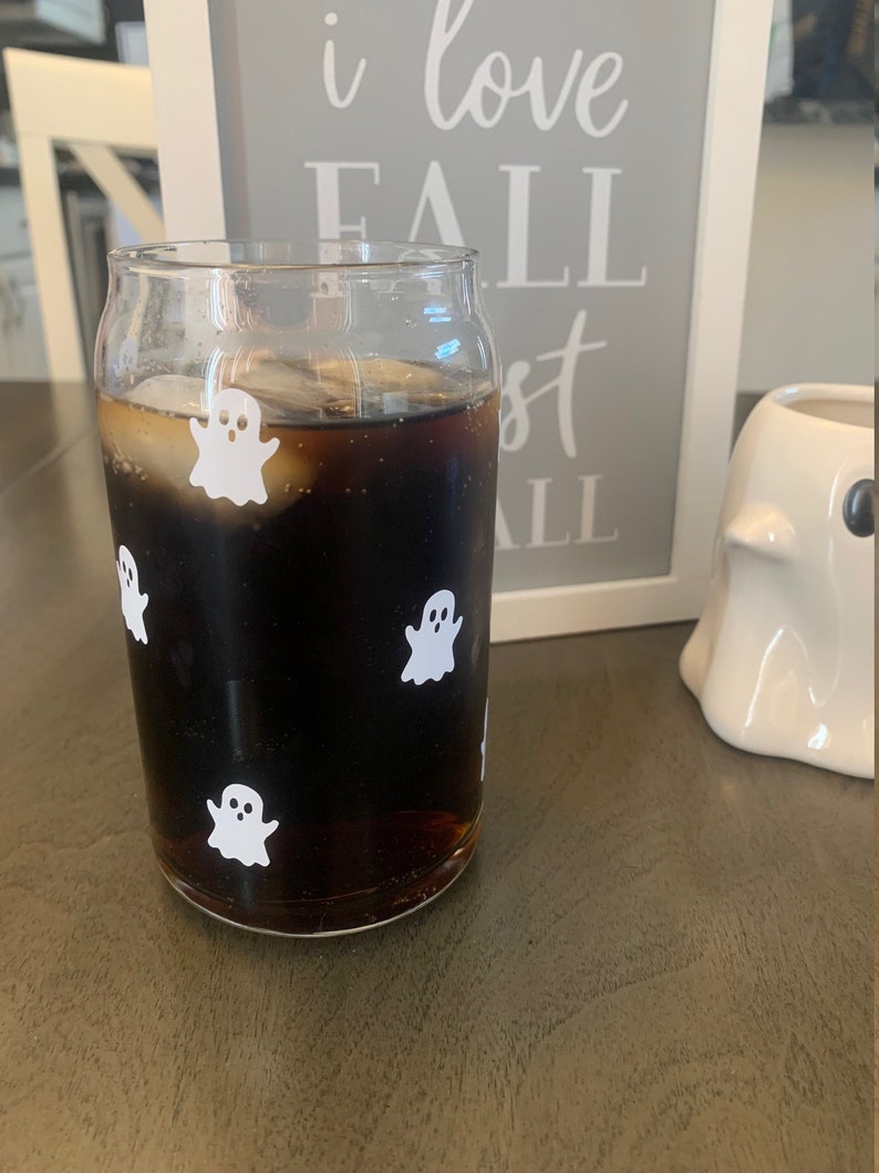 May include: A glass of cola with ice and ghost stickers on the side. The glass is sitting on a wooden table next to a white ghost figurine and a framed sign that says "I love fall most of all".