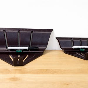 May include: Two dark brown miter saw guides with white and black markers. The guides are angled with slots and a ruler along the top edge. One guide is larger than the other. The guides are on a wooden surface.