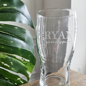May include: A clear pint glass etched with the name "RYAN" and the word "groomsman". The glass is on a wooden surface, with a large green plant visible in the background. The glass is suitable for serving beer or other beverages.