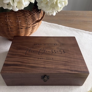 May include: A rectangular dark brown wooden box with a hinged lid and a small metal latch. The lid is engraved with the text RYAN & VICKI, The Chessers, and EST. 2019. The wood features a visible natural grain pattern throughout the surface.