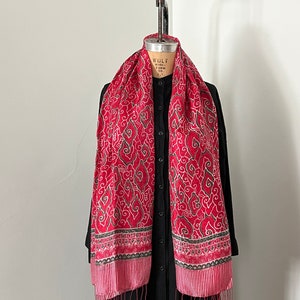 May include: Red silk scarf with a white and green floral pattern. The scarf has a fringe border and is displayed on a mannequin.