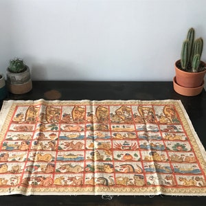 Op de afbeelding: Een vintage handgeschilderd stoffen rol die een reeks kleurrijke scènes in een rasterpatroon weergeeft. De rol ligt op een donker houten oppervlak met twee kleine potcactussen op de voorgrond.