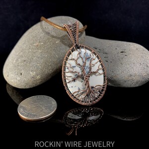 Copper wire tree of life wrapped around an oval white cabochon with small, black inclusions going through it. A brown waxed cord necklace runs through the bail. For size comparison, a US Quarter lays next to pendant. Coin is about 1.5 times smaller.