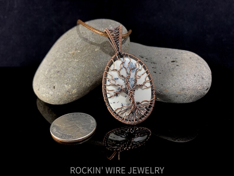 Copper wire tree of life wrapped around an oval white cabochon with small, black inclusions going through it. A brown waxed cord necklace runs through the bail. For size comparison, a US Quarter lays next to pendant. Coin is about 1.5 times smaller.