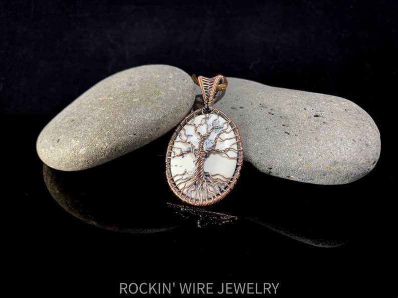 Copper wire tree of life wrapped around an oval white cabochon stone with small black, thin inclusions going through it. Copper is antiqued in order to show off the details of the wire weaving. A brown waxed cord necklace runs through the bail.