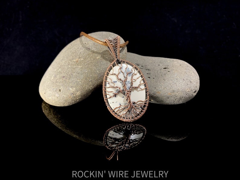 Copper wire tree of life wrapped around an oval white cabochon stone with small black, thin inclusions going through it. Copper is antiqued in order to show off the details of the wire weaving. A brown waxed cord necklace runs through the bail.