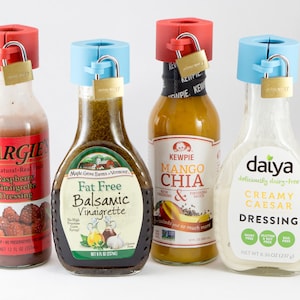 May include: Four bottles of salad dressing with colorful labels, each secured with a red or blue plastic cap and a small padlock. The dressings include raspberry vinaigrette, balsamic vinaigrette, mango chia, and creamy caesar.