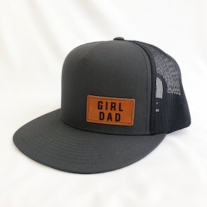 May include: A black and gray trucker hat with a brown leather patch that says "GIRL DAD" in black lettering.